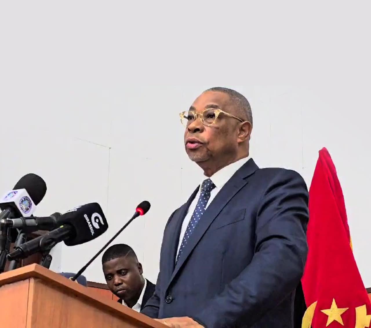 Eugénio Laborinho em pé num púlpito de madeira, discursando perante vários microfones de órgãos de comunicação social. Ele veste um fato azul escuro, gravata de pintas e óculos de armação clara. Ao fundo, vê-se parcialmente a bandeira de Angola.