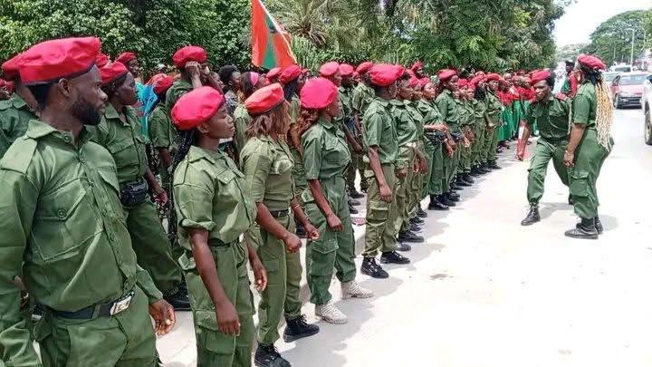 Fotografia de plano médio mostrando dezenas de jovens militantes da UNITA, homens e mulheres, formados em parada numa rua do Sumbe. Vestem uniformes verdes e boinas vermelhas. Ao fundo, vê-se uma bandeira da UNITA