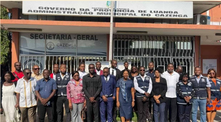 Fotografia de grupo em frente ao edifício da Administração Municipal do Cazenga, do Governo da Província de Luanda. Cerca de vinte jovens, homens e mulheres, estão posicionados em várias filas para uma foto oficial. Alguns vestem coletes azuis escuros com logótipos institucionais, enquanto outros usam trajes formais ou casuais. O letreiro superior identifica a "Secretaria-Geral" e a administração local.