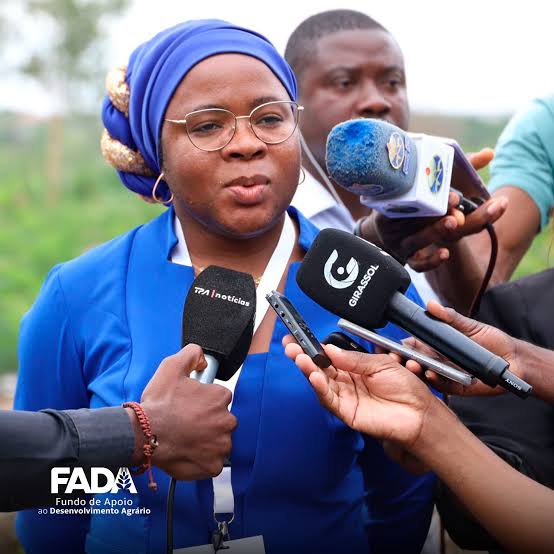 Mulher negra de óculos e turbante azul com detalhes em dourado, Felisbela Francisco, falando para vários microfones de jornalistas. Ela veste uma blusa azul royal. Em destaque, à frente, estão microfones da TPA Notícias e Girassol. No canto inferior esquerdo, lê-se o logótipo "FADA - Fundo de Apoio ao Desenvolvimento Agrário".