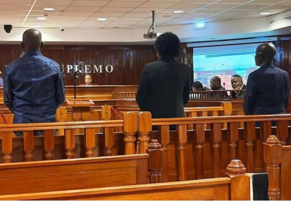 Fotografia do interior de uma sala de tribunal em Angola, vendo-se três pessoas de costas em primeiro plano perante o coletivo de juízes. Ao fundo, a inscrição 'Tribunal Supremo' na parede de madeira e um ecrã de projeção ativo. O ambiente é formal, com mobiliário em madeira envernizada e iluminação de escritório