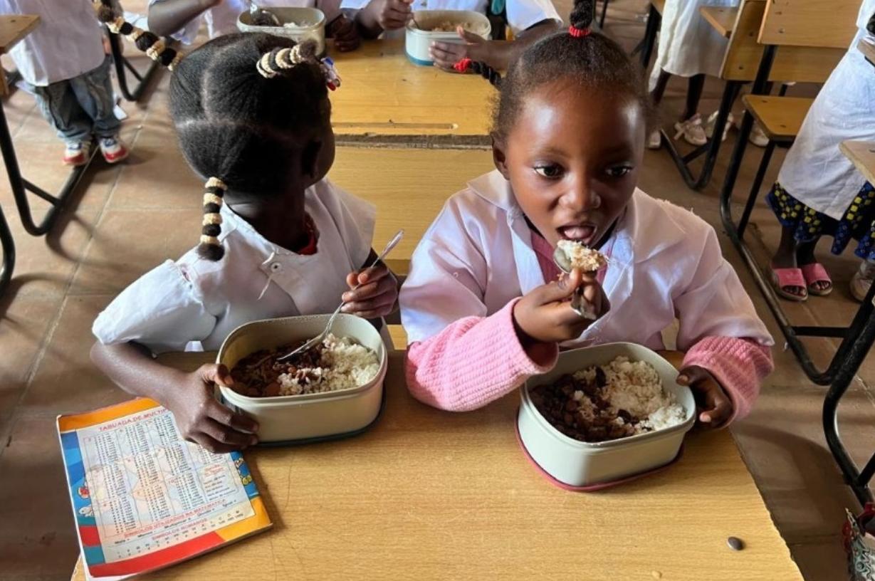 Crianças em sala de aula no Cuchi, Angola, consumindo merenda escolar precária apenas com arroz e feijão, sem água ou acompanhamentos.