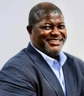 Retrato de plano médio de Pedro Inácio Velasco Galiano, um homem negro de meia-idade, sorrindo de forma leve e segura. Ele possui a cabeça raspada e uma barba curta e grisalha bem aparada. Veste um paletó azul escuro de corte clássico e uma camisa social azul claro sem gravata. O fundo é cinza neutro e limpo.