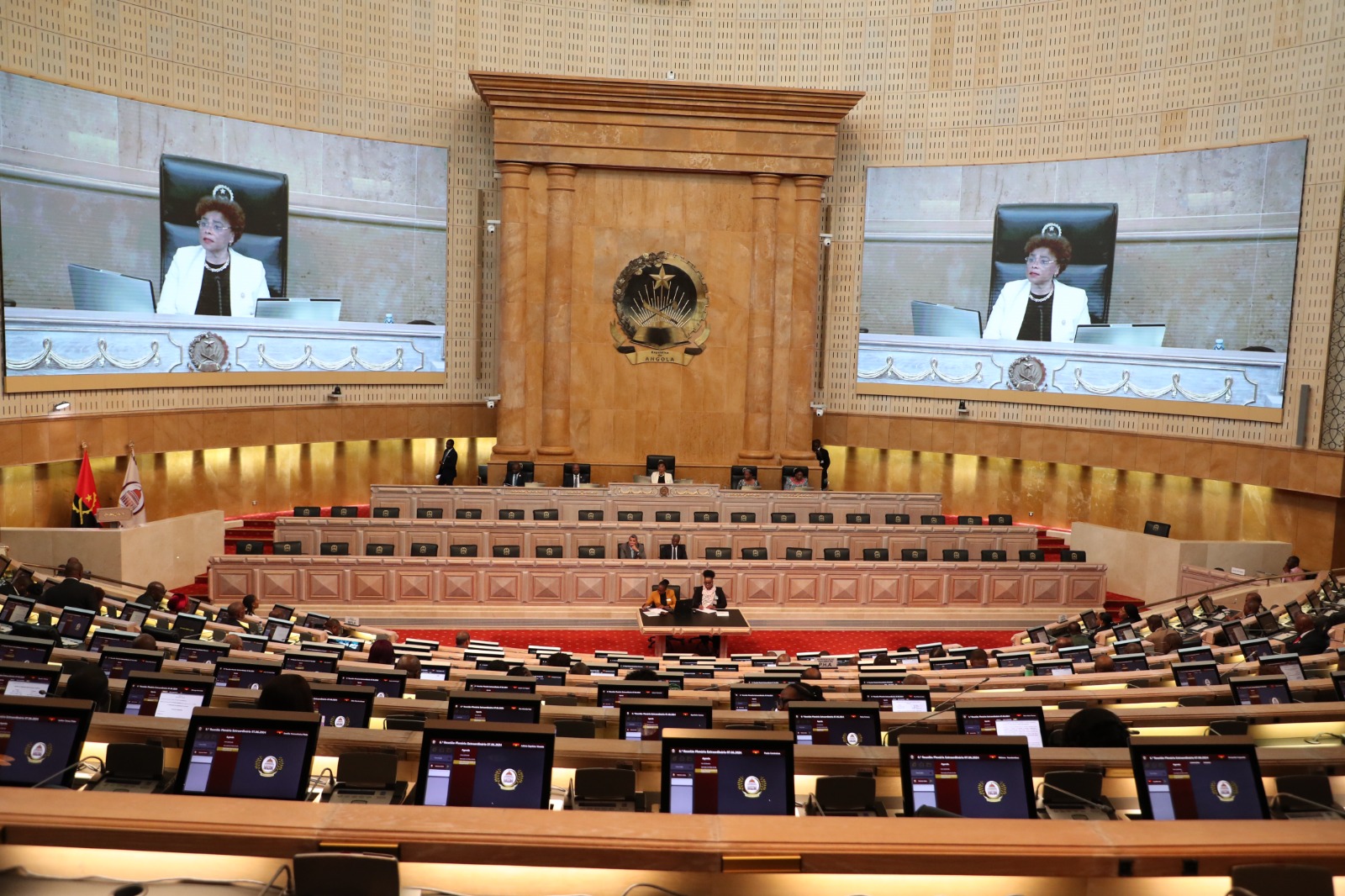 Fotografia panorâmica do interior da Assembleia Nacional de Angola durante uma sessão plenária. No centro, o emblema da República de Angola em relevo dourado sobre painéis de madeira. Dois grandes ecrãs laterais exibem a imagem da Presidente da Assembleia Nacional. As bancadas estão ocupadas por deputados e equipadas com monitores individuais.