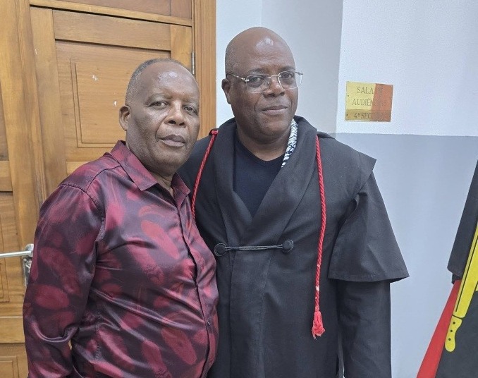 Imagem mostra David Mendes, à esquerda, e Robertinho, à direita, ambos sorrindo. David Mendes veste um terno preto e Robertinho veste uma camisa xadrez vermelha e preta. Eles estão em pé lado a lado, com David Mendes com o braço em volta do ombro de Robertinho.